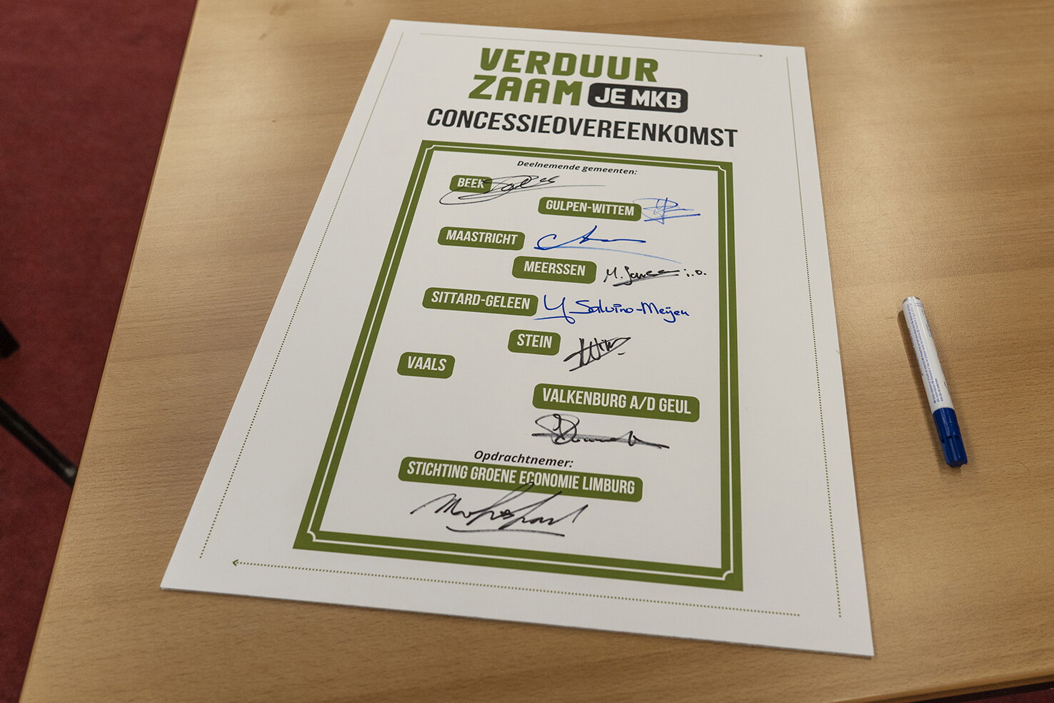 Ondertekende concessie met handtekeningen door gemeentes gemeentes Maastricht, Sittard-Geleen, Stein, Beek, Meerssen, Valkenburg aan de Geul, Vaals en Gulpen-Wittem VerduurzaamjeMKB Concessieovereenkomst staat op het papier. Papier is wit en gebruikte kleur voor tekst is groen. Het papier ligt op een tafel met naast het papier een witte pen met zwarte dop.