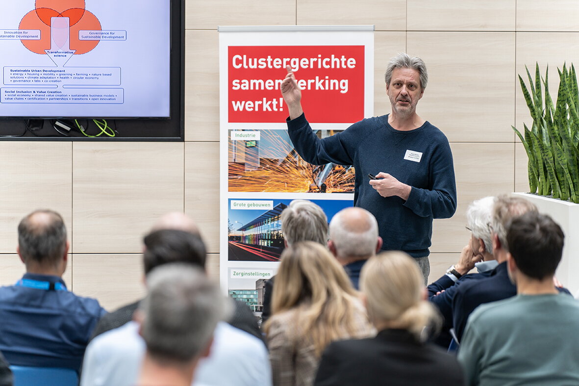 Spreker Frank Boons van de UM tijdens MEA Kennissessie 18 april 2024 - Frank spreekt tegen de groep en heeft zijn hand opgestoken. Naast hem hangt een presentatiescherm tegen de muur. Op de voorgrond zie je mannen en vrouwen zitten in de zaal. Je kijkt tegen hun rug aan. Het zijn elf personen. Achter Frank zie je ook nog een roll-up banner met in een rood vlak de tekst Clustergerichte samenwerking werkt!. Daaronder zie je twee foto's waarvan een van een gebouw en de ander waar maakindustrie aan het werk is.