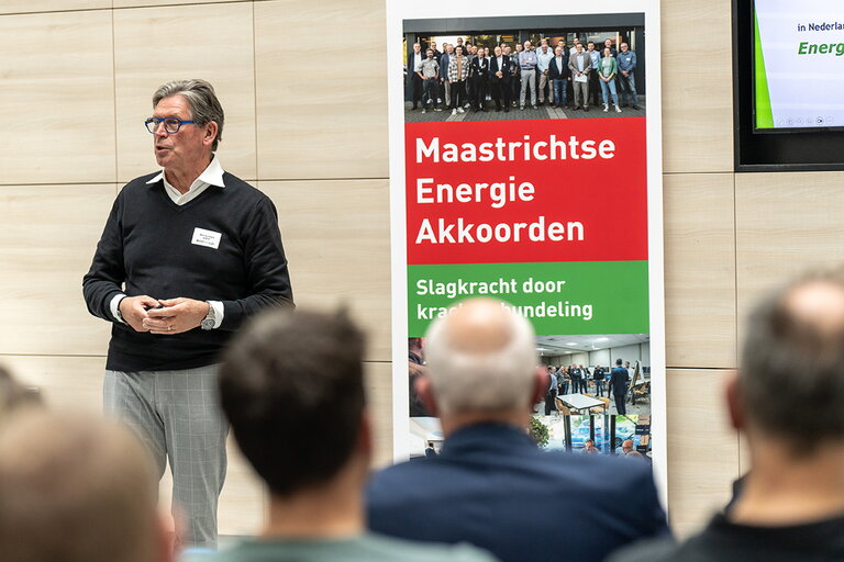 Spreker Wim Schilders bedrijventerrein Pannenweg in Nederweert tijdens MEA Kennissessie 18 april 2024. Wim staat voor de groep en op de voorgrond die je achterhoofden van een viertal mannen die in de zaal zitten. Naast Wim staat een rood/groene roll-up banner met beeldmateriaal. Op de roll-up banner staat in het rode vlak Maastrichtse Energie Akkoorden.