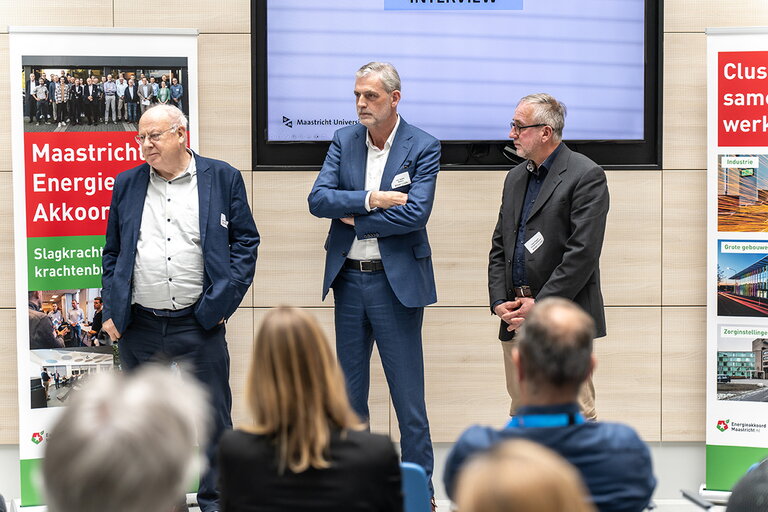 Sprekers John Aarts, Jan Janssen en Leon Kiggen tijdens MEA Kennissessie 18 april 2024. Ze staan naast elkaar voor de groep en luisteren naar iemand buiten beeld. Achter ze hangt een presentatiescherm tegen de muur en naast ze staat aan beide kanten een roll-up banner over energie akkoorden. Voorgrond zitten de aanwezigen die kijken naar de sprekers.